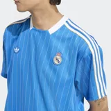 Remera de fútbol Adidas del Real Madrid, modelo Terrace Icons. Diseño celeste con finas rayas verticales blancas, cuello redondo blanco, mangas cortas con tres tiras blancas en los hombros, logo de Adidas y escudo del Real Madrid bordados en el pecho. Inspirada en la moda del fútbol de los años 80.