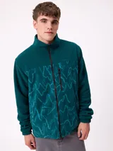 Campera reversible con cuello alto y cierre frontal. Un lado presenta tejido de polar con estampado geométrico en tonos verdes y bolsillo con cierre en el pecho, mientras que el otro lado es de nylon. Cuenta con puños elásticos y bolsillos laterales.