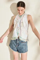 Top sin mangas de color blanco con estampado multicolor y detalles de volados desflecados, cuello mao y cierre frontal con botones.