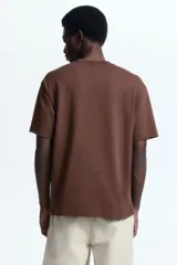 Remera de manga corta color marrón, con cuello redondo y corte holgado. Presenta un pequeño estampado en el pecho con la figura de una rana en color lila y la inscripción "ADAPTO OUTDOOR SUPPLIES".