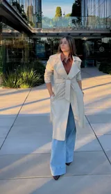 Trench coat de color beige, con diseño de doble botonadura, solapas amplias y cinturón ajustable en la cintura. Cuenta con mangas largas con trabillas en los puños y bolsillos laterales.
