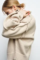 Jersey de punto color beige con cuello subido, manga larga y detalle de botones en los puños. Presenta costuras marcadas y acabados en rib.