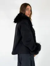 Chaqueta de textura gamuzada en color marrón, con solapas amplias y puños adornados con piel sintética de pelo largo en tono contrastante. Presenta un diseño de corte recto y estructura abierta.