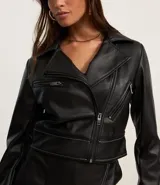 Campera biker cropped confeccionada en cuero sintético negro, con cierre cruzado con cremallera, cuello con solapa, pespuntes contrastantes y bolsillos con cremallera.