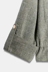 Chaqueta corta gris de tejido texturizado con cuello solapa, manga larga con trabilla, bolsillos delanteros con solapa y cierre frontal de botones metálicos.