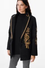 Blazer de textura suave en color crudo, con solapas clásicas y bordados florales en tonos tierra en el frente y las mangas. Presenta detalles de costura decorativa en los bordes de las solapas.