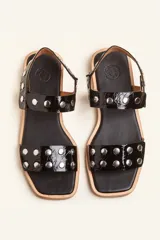 Sandalia baja de cuero vacuno color negro con acabado efecto charol texturizado, decorada con tachas metálicas plateadas en las tiras y la sujección del talón. Cuenta con suela plana de cuero.
