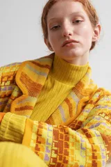 Saco largo medio con abertura frontal, bolsillos delanteros y cinto para ajustar. Presenta un diseño tejido con patrón geométrico en tonos amarillo, marrón y lila.