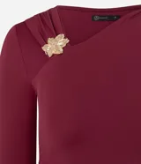 Body de poliamida color bordó, con diseño ajustado, mangas largas y escote asimétrico. Presenta detalles drapeados en la cintura y el hombro, junto con un adorno de flor metálica en el escote.