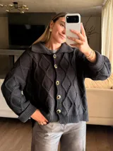 Saco tejido de punto con diseño de ochos y trenzas, color negro. Presenta cuello estilo camisero, cierre frontal con botones tipo joya y mangas holgadas con puños acanalados. Silueta oversize.