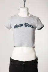 Remera de morley de manga corta, color gris, ajustada al cuerpo y elastizada, con bordado estilo college con la inscripción 'New York' en el pecho.