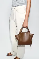 Bolso bandolera de formato city color marrón, con diseño estructurado, pespuntes decorativos, doble asa de mano y cremalleras laterales con tiradores colgantes.