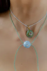 Gargantilla tipo bolo tie de tiento trenzado verde con dije esférico celeste con brillos incrustados.