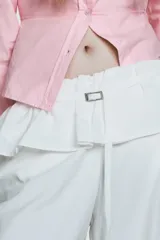 Pantalón blanco de nylon, tiro alto, con cintura elástica y detalle de sobrefalda o peplum superpuesto con hebilla metálica. Corte ancho y pierna recta.