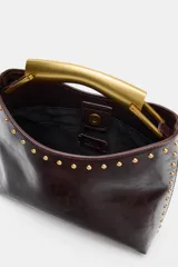 Bolso de mano de efecto acharolado color marrón oscuro, con detalle de tachas metálicas doradas en los bordes. Cuenta con asa rígida metálica dorada y cierre de botón imantado.