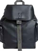 Mochila negra de estilo cargo con textura granulada, solapa superior con cierre de correa ajustable y hebilla metálica. Presenta un asa de mano superior y una correa textil gris con el logo "Calvin Klein" en relieve. Incluye un logo CK grabado en la parte inferior frontal.