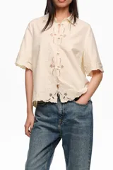 Blusa color beige, de manga corta, con cuello clásico, cierre frontal con botones revestidos y detalles de bordados calados en el frente, cuello, mangas y dobladillo.
