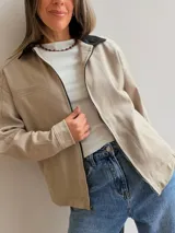 Campera de gabardina color beige con cierre frontal y cuello en contraste color negro. Presenta un diseño minimalista con bolsillos delanteros funcionales y calce cómodo.