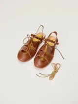 Sandalias planas tipo slingback de cuero marrón, con diseño delantero calado, cierre de cordones y tira trasera ajustable con hebilla.