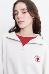 Buzo de felpa color crudo con cuello alto, cierre metálico y bordado de un corazón con rayos en el pecho.