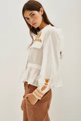 Camisa de algodón celeste con detalles de volados en los hombros y bordados en color negro en el pecho y las mangas. Presenta cierre frontal con botones y puños ajustados.