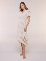 Vestido midi blanco de estilo romántico confeccionado en poliéster con elastano, con detalles de encaje floral. Cuenta con escote cruzado en V, mangas cortas acampanadas, cintura elastizada y falda evasé con terminación asimétrica en picos.