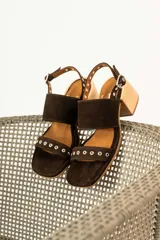 Sandalias de gamuza color marrón con taco cuadrado de madera. Presentan dos tiras anchas, una superior lisa y una inferior decorada con ojales metálicos plateados. Cuentan con cierre de pulsera con hebilla ajustable en el tobillo.