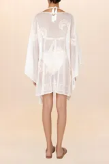 Salida de playa corta tipo poncho en color blanco marfil, confeccionada en algodón ligero y traslúcido. Presenta escote en V con lazo frontal ajustable terminado en borlas y detalles bordados en tono crema.