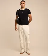 Remera estilo baby tee de color blanco, confeccionada en algodón, con cuello redondo y ribetes en color marrón en cuello y puños. Cuenta con un pequeño tag gomosa con el logotipo "Lucky" en el centro del pecho.