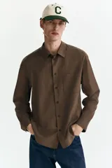 Camisa marrón de manga larga, confeccionada en sarga suave con cuello inglés, cierre frontal de botones, bolsillo en el pecho y bajo redondeado.