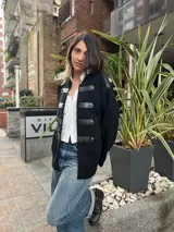 Campera estilo militar color negro con detalles de tiras de cuero sintético y botones metálicos en el frente.