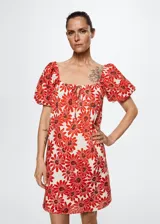 Vestido corto de algodón con estampado de margaritas rojas sobre fondo blanco. Presenta un diseño evasé, escote cuadrado con cierre de lazo ajustable, mangas cortas abullonadas y forro interior.