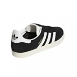 Championes urbanos Adidas modelo Gazelle, color negro con las icónicas tres tiras laterales y refuerzo en el talón en color blanco. Presenta el nombre del modelo en letras doradas en el lateral y suela de goma texturizada en color crema.