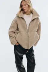 Campera de sherpa suave en color beige, de corte holgado con cuello elevado y cierre frontal bidireccional. Presenta bolsillos frontales tipo canguro y detalles de ribetes en tejido revestido.