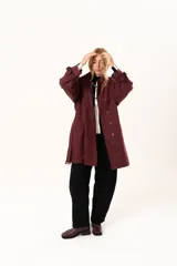 Trench de corte holgado color bordó, con cuello camisero, cierre frontal con botones y cinturón ajustable con hebilla. Presenta mangas largas con detalles de correas en los puños y bolsillos laterales.
