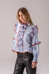 Camisa celeste con volados decorativos en los hombros, pecho, mangas y ruedo, destacados con ribetes en tono rojo. Presenta cierre frontal de botones, cuello alto y mangas largas abullonadas con puños ajustados.