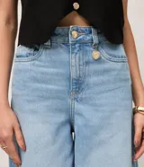 Pantalón de jean estilo pantalona con cintura alta, corte amplio desde el cuadril y cinco bolsillos funcionales. Presenta cierre frontal con botón y cremallera, además de un detalle de charm metálico en la cintura.