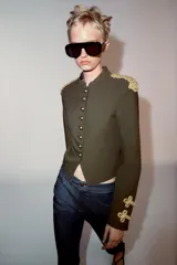 Cazadora militar de color verde oscuro, de cuello subido y manga larga. Cuenta con detalles de alamares y cordones dorados en los hombros y puños, y cierre frontal con botones dorados metálicos.