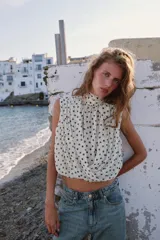 Blusa sin mangas blanca con estampado de lunares negros, cuello alto drapeado y nudo frontal en el bajo.