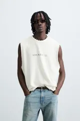 Camiseta sin mangas (manga sisa) de punto color crema, corte relaxed fit, con cuello redondo y acabados irregulares en las sisas. Presenta un texto estampado en el pecho que dice "SERIAL N. 8763/ 7.3Z".