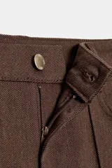 Pantalón de tiro alto color marrón oscuro, con corte flare (campana) y nervio marcado en el frente de ambas piernas. Presenta bolsillos delanteros y bolsillos de plastrón en la espalda.