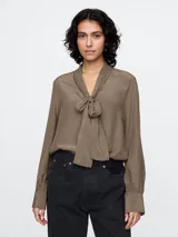 Blusa de satén color marrón topo, con cuello tipo lazo o moña, mangas largas con puños abotonados y corte fluido.