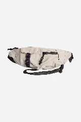 Riñonera unisex de diseño urbano en color beige claro, confeccionada en nylon. Presenta múltiples bolsillos frontales con cierres y tiradores negros, incluyendo un bolsillo con tapa y hebilla de ajuste negra. La correa es de nylon oscuro con texto repetido.