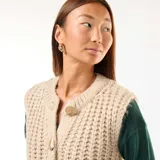 Chaleco de punto beige con textura de ochos, sin mangas, con cuello redondo y cierre frontal con botones grandes dorados.