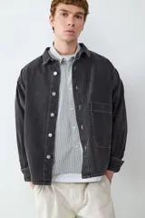 Sobrecamisa de denim en color negro lavado, con cierre frontal de botones metálicos, cuello clásico y un bolsillo de parche en el pecho. Presenta costuras a la vista y puños abotonados.
