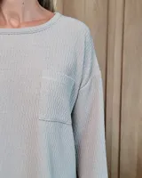 Remerón de textura acanalada en color gris claro, con cuello redondo, mangas largas y bolsillo frontal en el pecho. Presenta un diseño holgado con corte asimétrico, siendo más largo en la parte posterior.