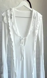 Vestido largo de microtul blanco, con escote en V profundo y mangas largas acampanadas. Presenta aberturas laterales en la falda y cierre con lazos en el frente. Es una prenda transparente.