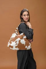 Bolso tipo hobo de gran capacidad con estampado animal print de vaca en tonos marrón y blanco. Presenta una etiqueta de cuero con el logo de la marca en el frente y un diseño de caída relajada.