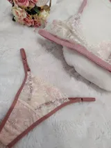 Conjunto de lencería compuesto por bralette y tanga regulable, confeccionado en encaje color champagne con detalles florales bordados en hilo rosado y terminaciones en cinta color rosa viejo.