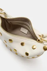 Bolso de hombro de diseño saca multiposición color blanco, decorado con tachas metálicas doradas y aros metálicos que conectan el cuerpo con la manija. Cuenta con cierre de cremallera metálica y bolsillo interior.